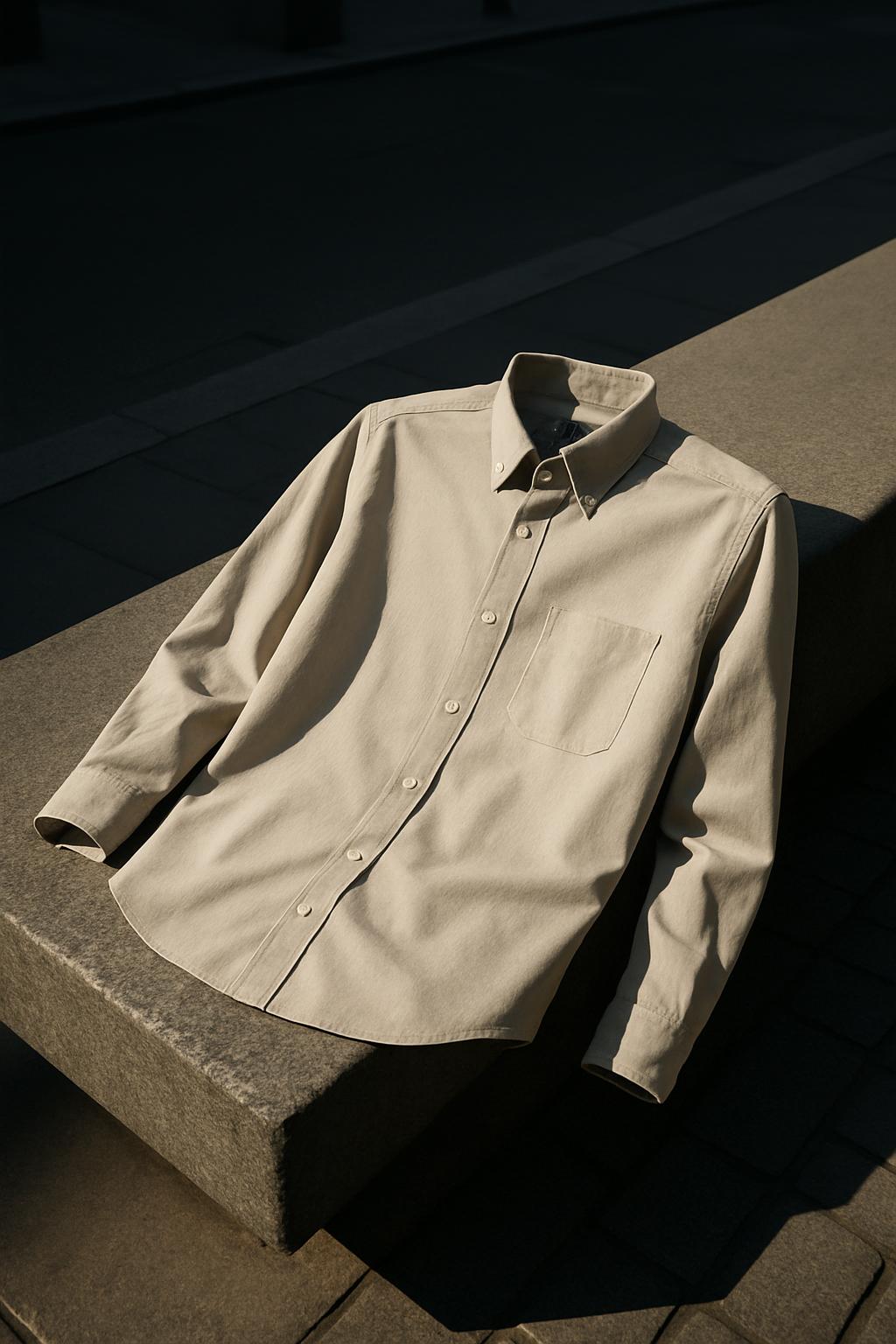 An unbuttoned, long-sleeved men's beige dress or casual shirt laid flat atop a stone bench and partially cast in a shadow,...